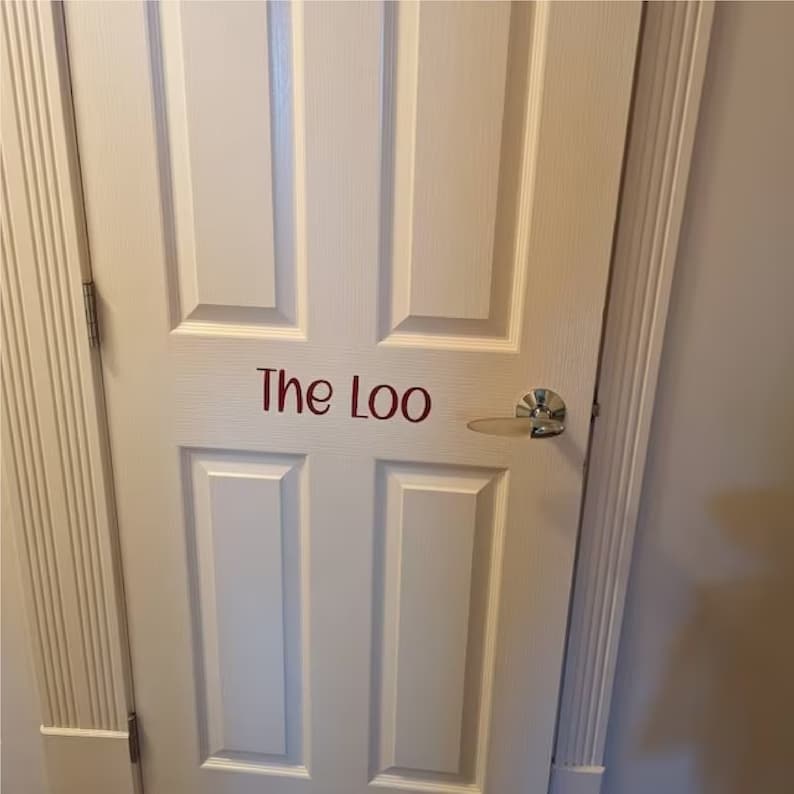 K&ouml;nnte beinhalten: Wei&szlig;e T&uuml;r mit einem silbernen T&uuml;rgriff und den Worten "The Loo" in roten Buchstaben.