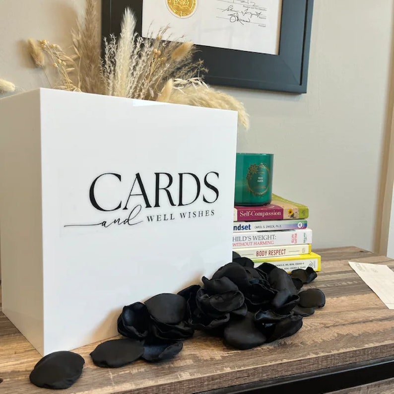 May include: White acrylic box with black text that reads "CARDS and WELL WISHES". The box is on a wooden table with black silk flower petals and a stack of books.