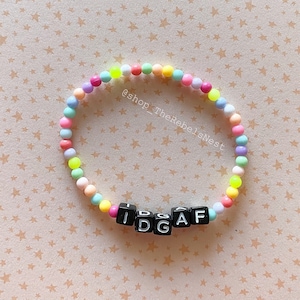 May include: A colorful beaded bracelet with the letters "IDGAF" spelled out in black letter beads. The bracelet is on a background of small pink stars.