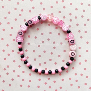 Puede incluir: Pulsera de cuentas rosa y negras con las letras "YOU GO GLEN COCO" deletreadas. La pulsera está sobre un fondo rosa con lunares.