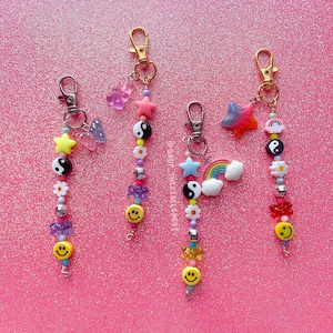 May include: Four beaded keychains with gold-colored clasps. Each keychain features a unique design with colorful beads, smiley faces, stars, yin-yang symbols, and other charms. The background is a sparkly pink surface.