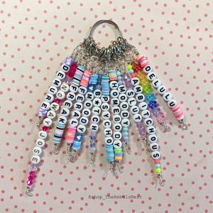 May include: A set of 12 colorful beaded keychains with different words spelled out on each one. The words include "Delulu", "Ambitcho", "Slut", "Moody", "Homebody", "Psycho", "Btch", "Feral", "Freak", "Demure", "Noway", and "Jose".