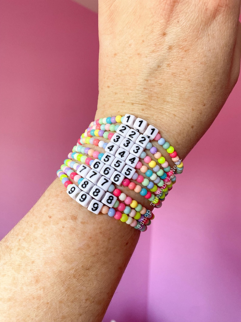 May include: A stack of colorful beaded bracelets with white square beads that have numbers 1 through 9 printed on them. The bracelets are on a person's wrist.