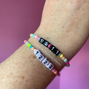 May include: Two colorful beaded bracelets with letter beads spelling out "1111" and "1222". The bracelets are on a person's wrist. The text "@shop_TheRebelsNest" is visible on the image.
