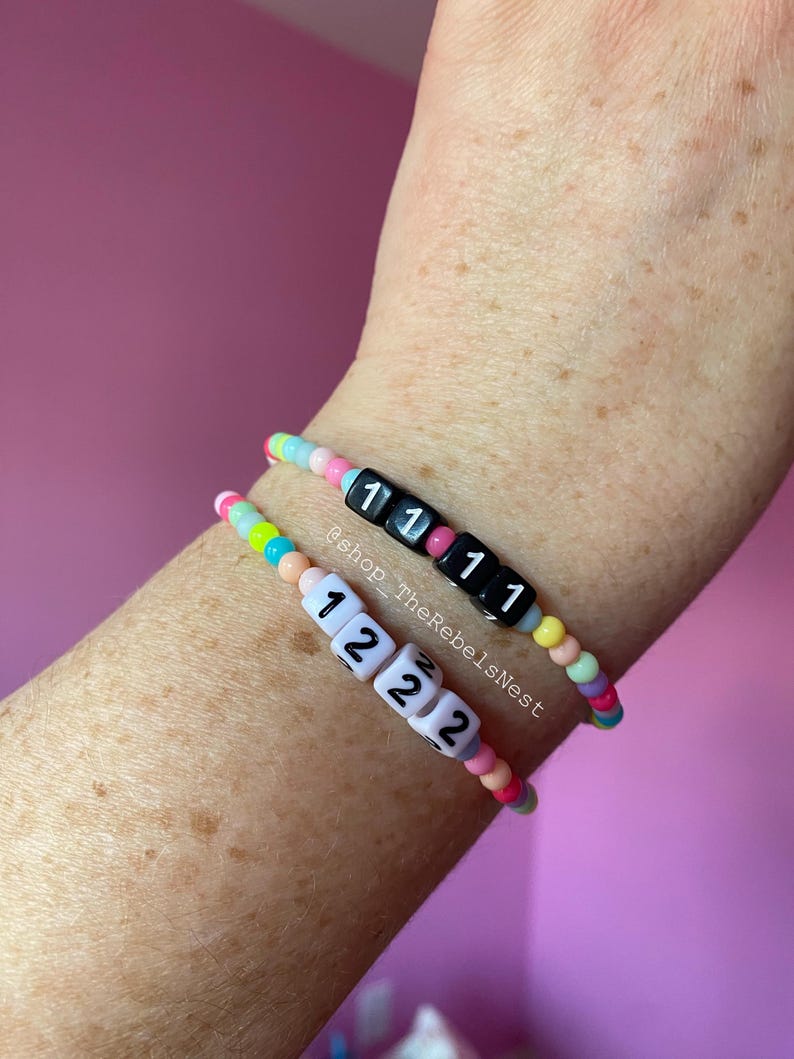 May include: Two colorful beaded bracelets with letter beads spelling out "1111" and "1222". The bracelets are on a person's wrist. The text "@shop_TheRebelsNest" is visible on the image.