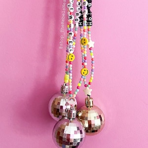 May include: Three hanging disco ball ornaments attached to colorful beaded strands. The beads include yellow smiley faces, white stars, and black letter beads spelling out phrases. Each silver mirrored sphere features a metallic cap and a small rainbow charm.
