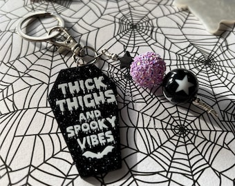May include: A Halloween-themed keyring featuring a black coffin-shaped charm with the text "THICK THIGHS AND SPOOKY VIBES" in white. It includes a purple glitter bead, a black bead with a white star, and a small black star charm. The background is a spiderweb.