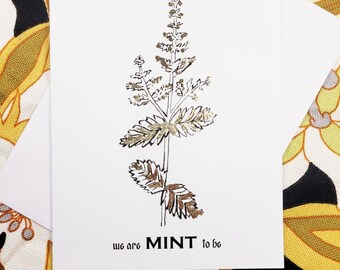 Op de afbeelding: Een witte kaart met een gouden folie-illustratie van een muntplant. De tekst "we are MINT to be" is onder de plant gedrukt.