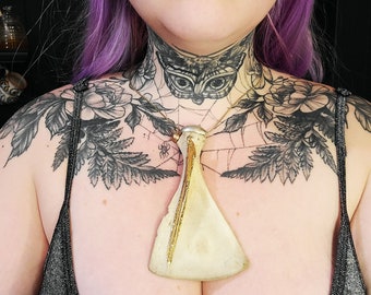 Op de afbeelding: Een persoon met paars haar en meerdere tatoeages draagt een ketting met een grote, witte, keramische hanger in de vorm van een bijl. De hanger heeft gouden accenten en hangt aan een dun, zwart koord.