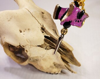 Op de afbeelding: Een paarse en gouden bedel met zwarte accenten, in de vorm van een klein wezen met hoorns, hangend aan een gouden ketting met een zwarte kraal en een zilveren pin.