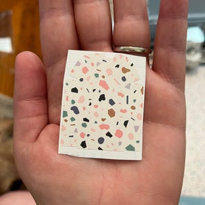 May include: A white sticker with a terrazzo pattern. The pattern features various shapes and colors, including pink, brown, green, blue, and black.
