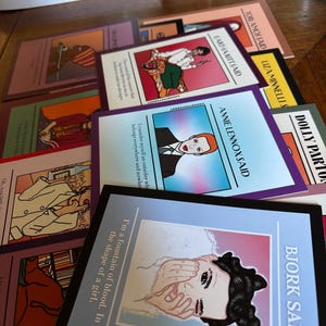 May include: A collection of colorful art cards featuring illustrations and quotes from various artists. The cards have a variety of designs, including portraits and text. The cards are arranged on a wooden surface.