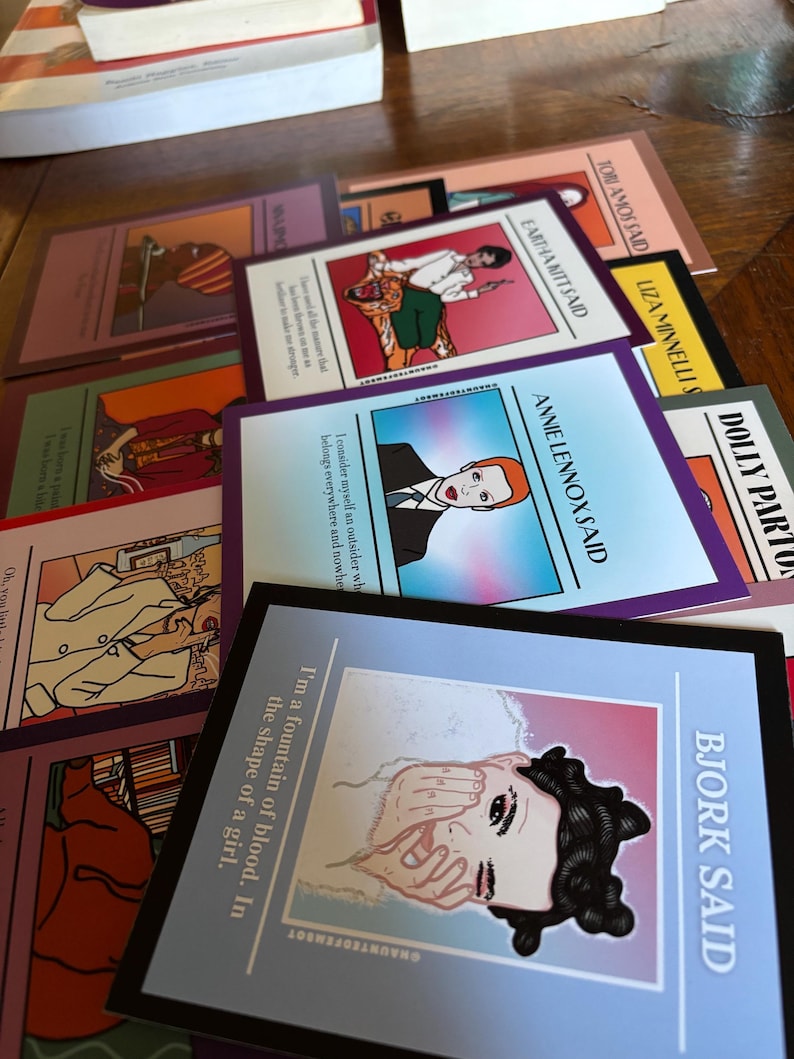 May include: A collection of colorful art cards featuring illustrations and quotes from various artists. The cards have a variety of designs, including portraits and text. The cards are arranged on a wooden surface.