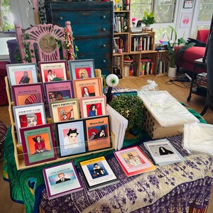 May include: A display of colorful books with illustrations and text, including titles like "Grace Jones Laid" and "Yoko Ono Said." The books are arranged on a wooden shelf and a table with a patterned cloth, showcasing a variety of artistic covers.