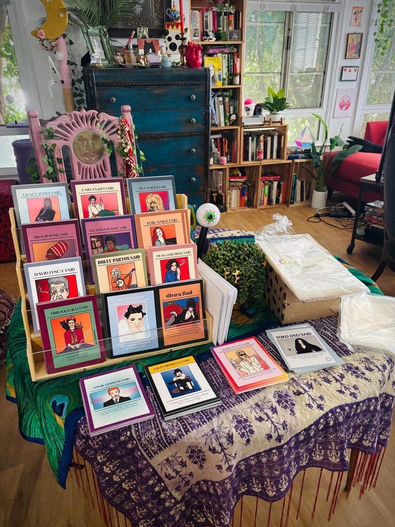 May include: A display of colorful books with illustrations and text, including titles like "Grace Jones Laid" and "Yoko Ono Said." The books are arranged on a wooden shelf and a table with a patterned cloth, showcasing a variety of artistic covers.