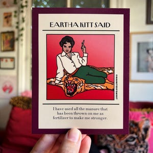 May include: A rectangular art print with a burgundy border features an illustration of Eartha Kitt. She is depicted in a white blouse and green pants, reclining on a tiger rug. The text reads "EARTHA KITT SAID" and a quote about resilience.