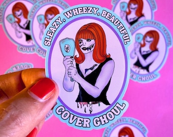 May include: A sticker with a cartoon illustration of a woman with red hair and a black dress. She is holding a mirror and looking at herself. The sticker is round and has a blue and purple border. The text on the sticker reads "Sleazy Wheezy, Beautiful Cover Ghoul".