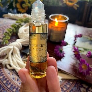 May include: A roll-on bottle containing amber liquid, with a label reading "Desert Moon." The label has a gold border and black text. The bottle is held in a hand, with a lit candle and flowers in the background.