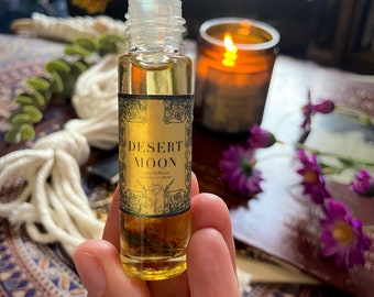 Pode incluir: Um frasco roll-on de vidro transparente preenchido com líquido âmbar, rotulado "Desert Moon". O rótulo tem uma borda dourada e texto preto. O frasco é segurado em uma mão, com uma vela acesa e flores ao fundo.