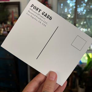 May include: A white postcard with black text that reads "POST CARD" and "@HauntedFembot" and "Haunted Fembot LLC. Los Angeles, CA, USA". The postcard has a black vertical line and a square for a stamp.