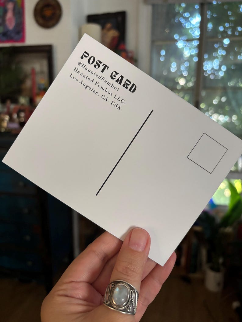 May include: A white postcard with black text that reads "POST CARD" and "@HauntedFembot" and "Haunted Fembot LLC. Los Angeles, CA, USA". The postcard has a black vertical line and a square for a stamp.