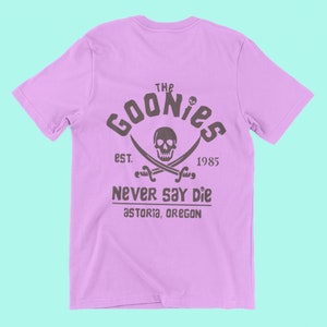 Op de afbeelding: Een lichtpaarse t-shirt met een zwart-witte grafische print van een schedel en gekruiste beenderen met de tekst "The Goonies Est. 1985 Never Say Die Astoria, Oregon"