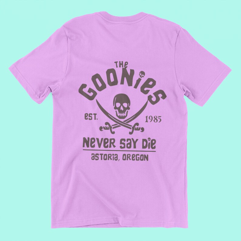 Op de afbeelding: Een lichtpaarse t-shirt met een zwart-witte grafische print van een schedel en gekruiste beenderen met de tekst "The Goonies Est. 1985 Never Say Die Astoria, Oregon"