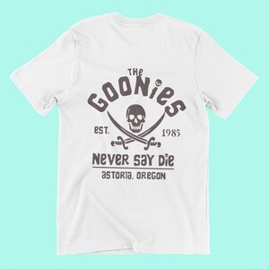 Op de afbeelding: Wit T-shirt met een bruine grafische print van een schedel en gekruiste beenderen met de tekst "The Goonies Est. 1985 Never Say Die Astoria, Oregon"