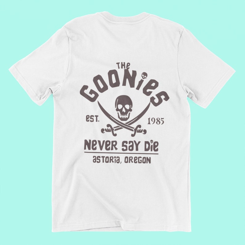 Op de afbeelding: Wit T-shirt met een bruine grafische print van een schedel en gekruiste beenderen met de tekst "The Goonies Est. 1985 Never Say Die Astoria, Oregon"