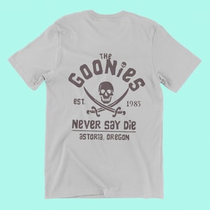 Op de afbeelding: Een grijs T-shirt met een zwart-witte grafische print van een schedel met gekruiste beenderen met de tekst "The Goonies Est. 1985 Never Say Die Astoria, Oregon"