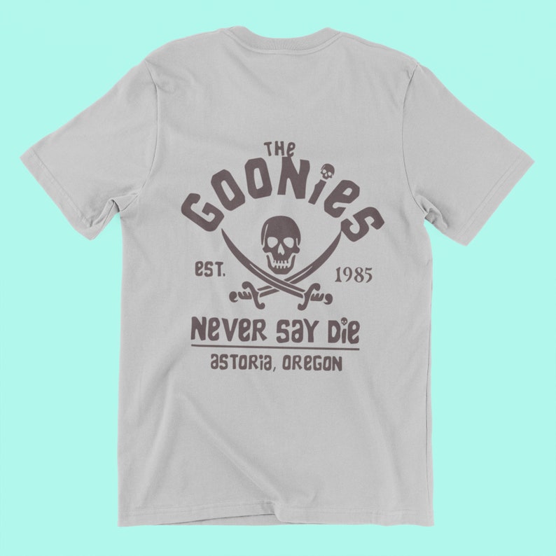 Op de afbeelding: Een grijs T-shirt met een zwart-witte grafische print van een schedel met gekruiste beenderen met de tekst "The Goonies Est. 1985 Never Say Die Astoria, Oregon"