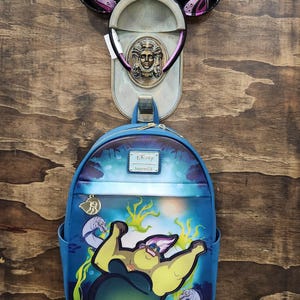 May include: A blue Disney Loungefly backpack featuring Ursula from The Little Mermaid. The backpack has adjustable straps and a top handle. Above the backpack is a decorative wall hanging with Mickey Mouse ears and a metal plaque with a face.