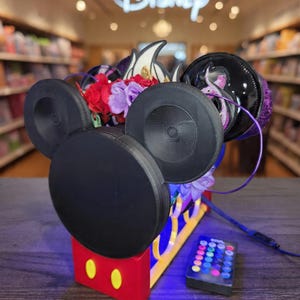 Puede incluir: Un auricular negro con forma de Mickey Mouse con detalles en rojo, amarillo y azul. El auricular presenta grandes auriculares negros y un arreglo floral decorativo. Un mando a distancia con botones coloridos está cerca. El logotipo de Disney es visible en el fondo.