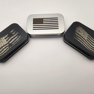 Puede incluir: Tres pequeñas latas rectangulares de metal con diferentes diseños de la bandera estadounidense. La lata superior es plateada con una bandera estadounidense en blanco y negro. Las dos latas inferiores son negras con un diseño de bandera estadounidense desgastado en marrón y blanco.