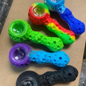 May include: An assortment of colorful silicone pipes. The pipes feature a honeycomb pattern and a bee design. Colors include green, red, yellow, blue, purple, and black. Each pipe has a glass bowl.