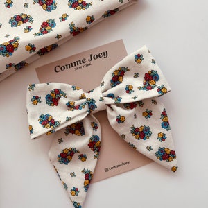 May include: A large white bow with a floral pattern in shades of red, yellow, blue, and green. The bow is made of fabric and has a pointed shape at the bottom. The bow is on a beige background with the text "Comme Joey NEW YORK" and "@commejoey" printed on it.