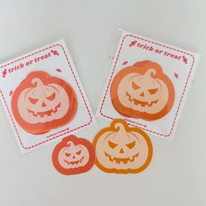 May include: Two clear plastic packages containing stacks of orange pumpkin-shaped sticky notes, each with a red dashed border and the text trick or treat. Two loose pumpkin sticky notes in different sizes are placed in front of the packages.