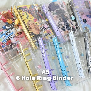 May include: A collection of A5 6-hole ring binders in various colors including pink, yellow, purple, blue, white, and black. The binders feature clear plastic covers decorated with diverse collage-style graphics, stickers, and attached charms.
