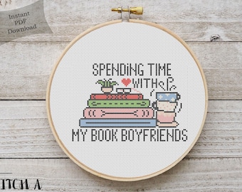 May include: A white embroidery hoop with the text "Spending time with my book boyfriends" in black. The text is surrounded by a stack of books, a potted plant, and a cup of tea.