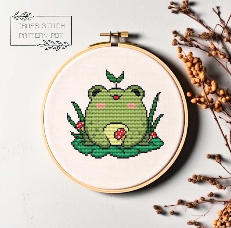 May include: A cross stitch pattern of a green frog sitting on a lily pad with red mushrooms. The frog has a small green leaf on its head. The pattern is in a hoop.