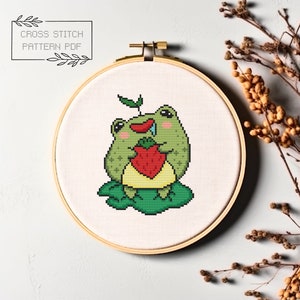 May include: A cross stitch pattern featuring a green frog holding a red strawberry. The frog is sitting on a green leaf. The pattern is in a hoop.