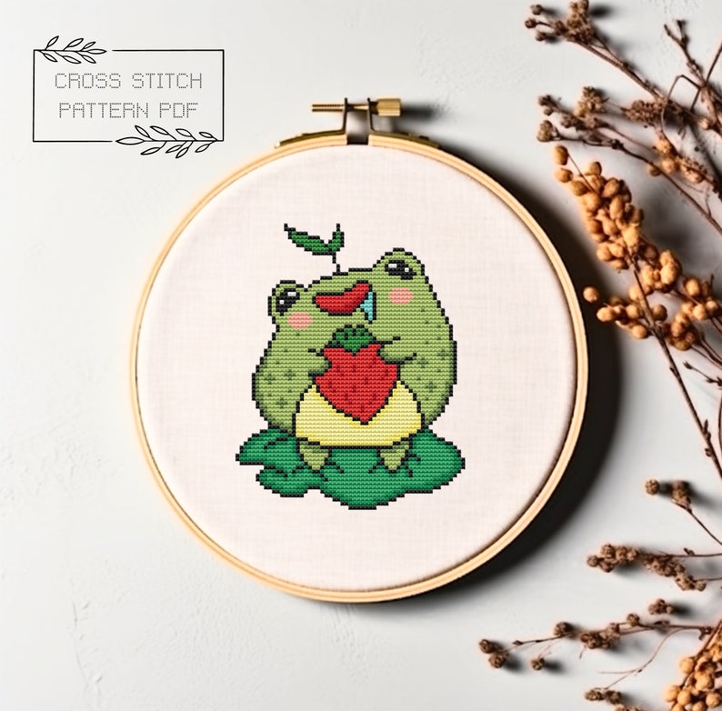 May include: A cross stitch pattern featuring a green frog holding a red strawberry. The frog is sitting on a green leaf. The pattern is in a hoop.