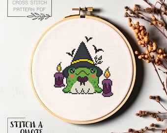 May include: Cross stitch pattern PDF of a green frog wearing a witch's hat with a black brim and yellow band. The frog is holding a purple bow tie and is surrounded by two purple candles with flames.