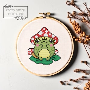May include: Cross stitch pattern PDF of a green frog wearing a red and white mushroom hat, sitting on a green leaf.