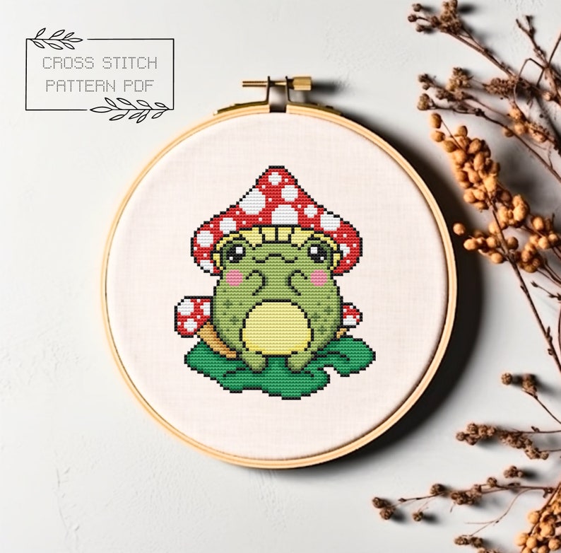 May include: Cross stitch pattern PDF of a green frog wearing a red and white mushroom hat, sitting on a green leaf.