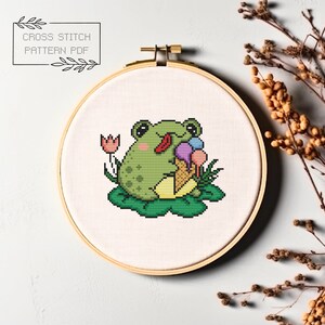 May include: Cross stitch pattern PDF of a green frog sitting on a lily pad, eating ice cream with a pink, purple, and blue scoop. The frog has a red tongue sticking out. There is a pink flower behind the frog.