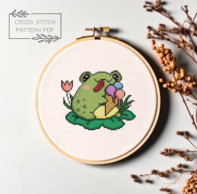 May include: Cross stitch pattern PDF of a green frog sitting on a lily pad, eating ice cream with a pink, purple, and blue scoop. The frog has a red tongue sticking out. There is a pink flower behind the frog.