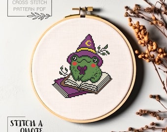 May include: A cross stitch pattern featuring a green frog wearing a purple wizard hat and reading a book. The pattern is displayed in a wooden embroidery hoop.