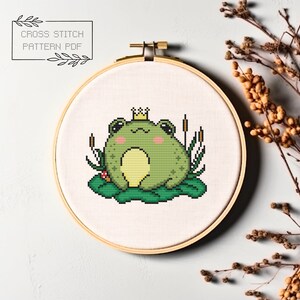 May include: Cross stitch pattern of a green frog wearing a gold crown, sitting on a lily pad with reeds in the background. The pattern is displayed in a wooden embroidery hoop.