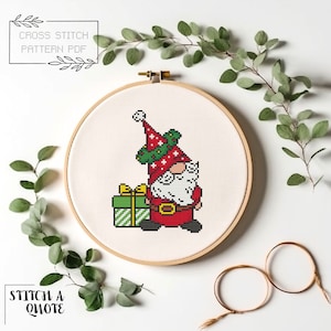 May include: A cross stitch pattern of a Christmas gnome holding a green and white striped gift box. The gnome is wearing a red hat with a green wreath and a white beard. The pattern is displayed in a wooden embroidery hoop.